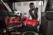 A person places a red Craftsman toolbox in the back of a vehicle, which is filled with various red and black Craftsman tool bags and fishing equipment. The scene appears to be inside a garage or workshop with windows in the background.