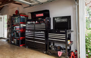 A garage interior showing multiple CRAFTSMAN branded black tool storage units with drawers and compartments, filled with tools and accessories. To the left, there are red and gray utility shelves holding storage bins and other items. The scene is organized, highlighting CRAFTSMAN tool chests and cabinets.