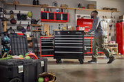 A person pushing a large black rolling tool chest in a garage workshop. The background shows several red tool cabinets, shelves with storage boxes, and various tools and equipment organized neatly. There are open black storage bins filled with sports equipment and other items on the floor.