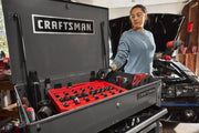 A CRAFTSMAN-branded tool chest is open, showing various tools organized in red trays. A person wearing gloves holds a power