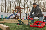 A person is kneeling on the grass using power tools to assemble wooden playground equipment. A red tool kit with various sockets and tools is open in the foreground. In the background, a child is on a swing and another person is standing nearby. The setting is an outdoor yard with a play structure and a white fence.